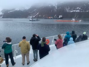 Photo of Valdez residents standing on the deck of the tour boat. They are watching several fishing boats practice pulling oil spill collection boom in the distance.