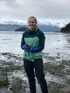 Photo of Mia holding a small crab discovered while setting traps for invasive European green crab. The crab she is holding is native to Alaska. 