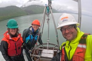 Three people stand in front of equipment used to collect sediment from the bottom of the sea floor.