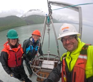 Three people stand in front of equipment used to collect sediment from the bottom of the sea floor.