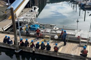 An instructor stand before a group of learners explaining details about a piece of oil spill response equipment. The equipment is a metal boat with a hollow interior that is used to hold the oil and water mix that is collected during an oil spill response. 