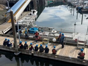 An instructor stand before a group of learners explaining details about a piece of oil spill response equipment. The equipment is a metal boat with a hollow interior that is used to hold the oil and water mix that is collected during an oil spill response. 