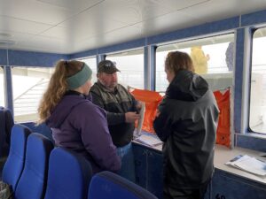 This photo shows three people inside the cabin of the tour boat discussing a piece of an oil skimmer. The piece is a round disc coated in a fuzzy material.