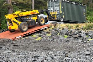 A large forklift carefully places a new conex on a concrete foundation. 